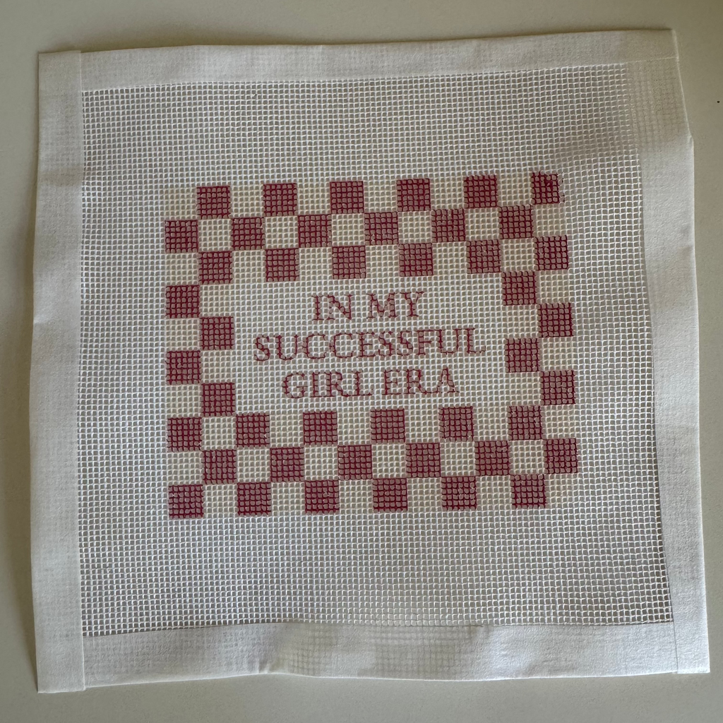 Cross-stitched fabric with red and white checkered pattern and text on a beige background with " In My Successful Girl Era" Saying