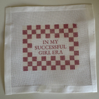Cross-stitched fabric with red and white checkered pattern and text on a beige background with " In My Successful Girl Era" Saying