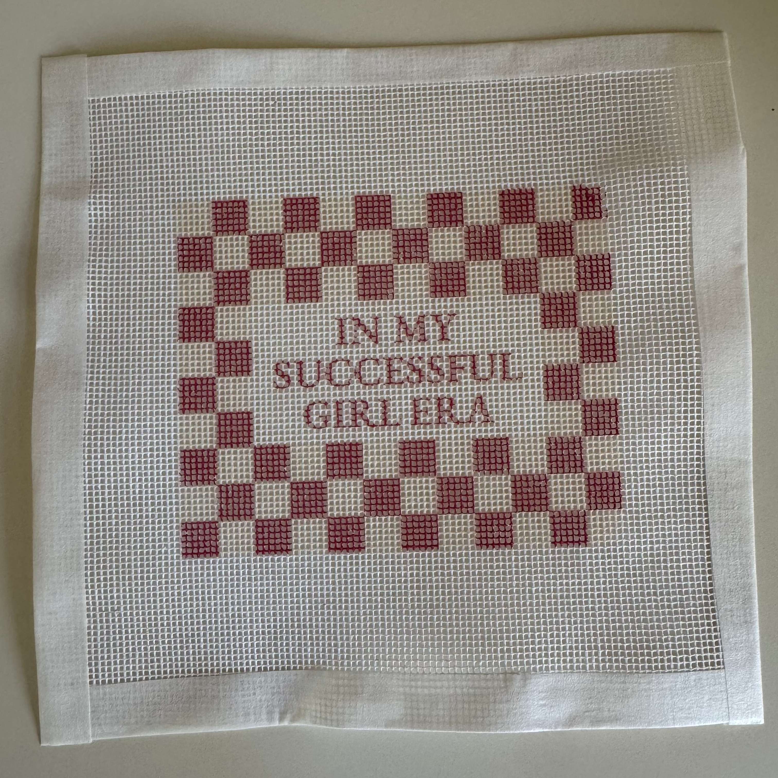 Cross-stitched fabric with red and white checkered pattern and text on a beige background with " In My Successful Girl Era" Saying