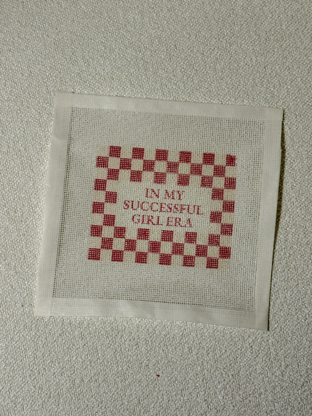 Cross-stitched fabric with red and white checkered pattern and text on a beige background with " In My Successful Girl Era" Saying