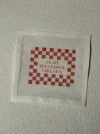 Cross-stitched fabric with red and white checkered pattern and text on a beige background with " In My Successful Girl Era" Saying