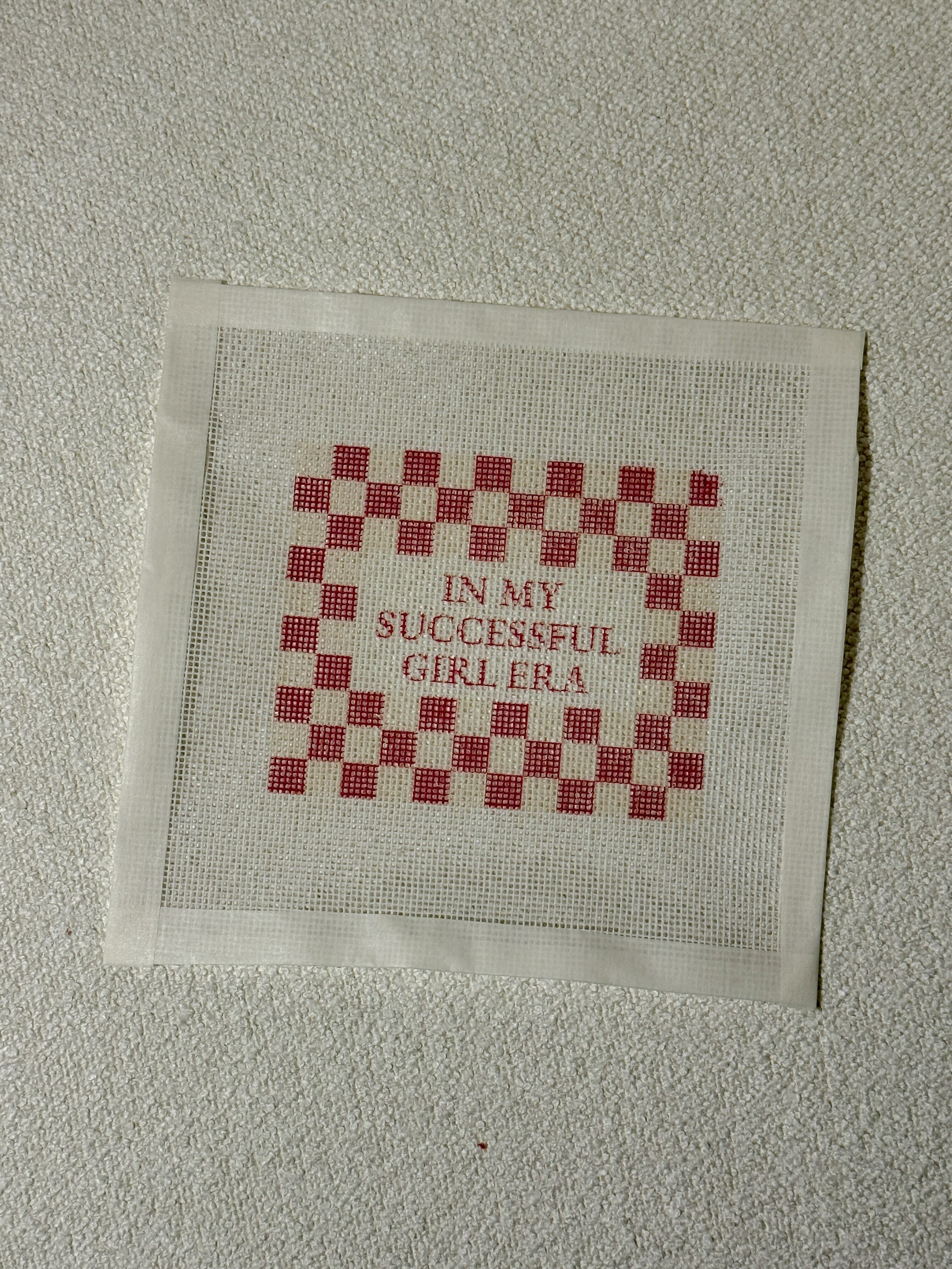 Cross-stitched fabric with red and white checkered pattern and text on a beige background with " In My Successful Girl Era" Saying